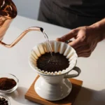 A barista pouring hot water from a gooseneck kettle over freshly ground coffee in a pour-over setup, creating a rich, aromatic brew.